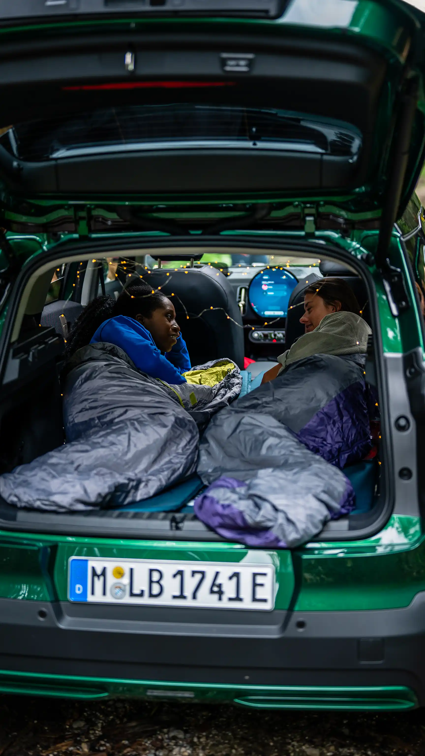 Two people lying in sleeping bags in the back of a MINI Countryman in Racing Green IV with the tailgate open.
